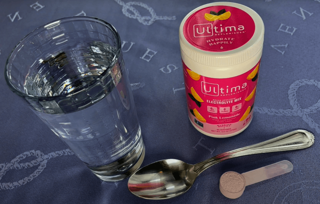 Ultima Replenisher electrolyte powder, pink lemonade flavor, with water and measuring scoop.